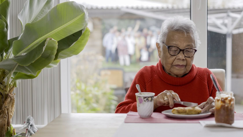 Gezellige oudere vrouw eet ontbijt met modern servies aan tafel in huis, grote plant op de voorgrond, rustige ochtend sfeer, overzicht van familie of bewoners, huiselijke setting, geschikt voor thema wellness, familie, comfort.