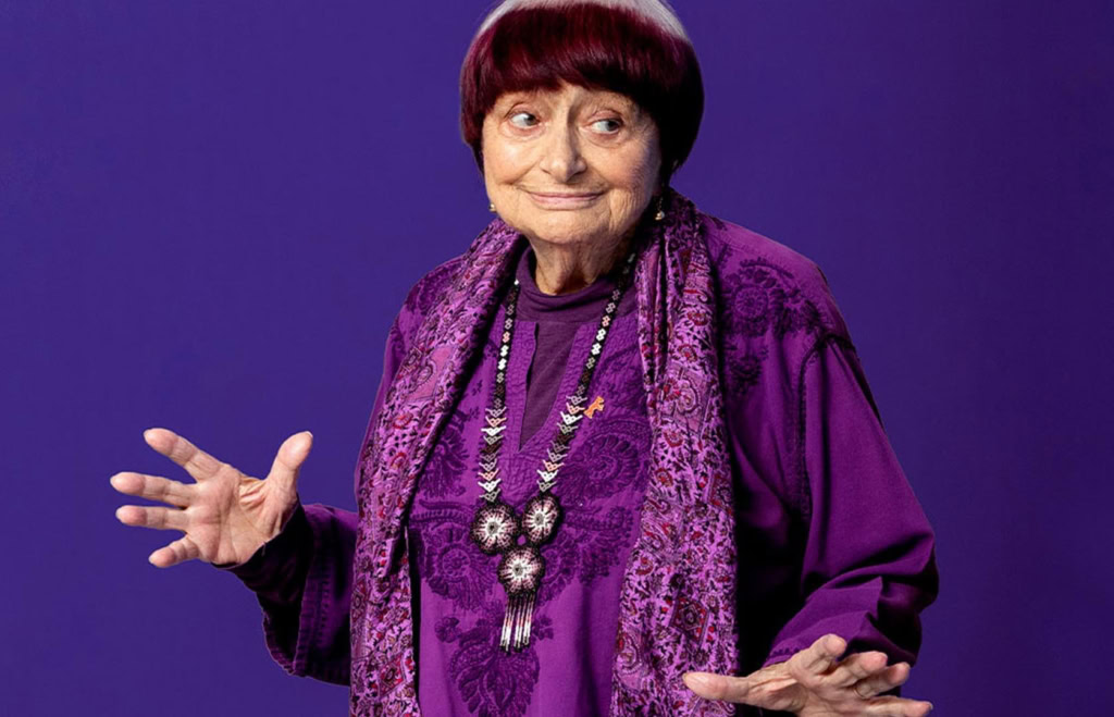 Kleine oude vrouw in een paarse outfit met sieraden, glimlachend en gebaren makend, op een effen paarse achtergrond.