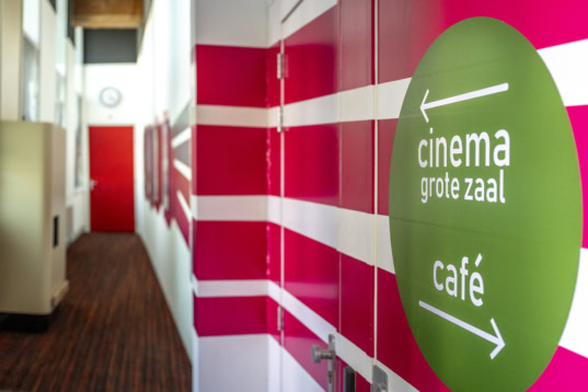 Gezellige ingang corridor van Cinema Middelburg met een opvallend groen signaalbord naar de grote zaal en café, kleurrijke muurdesign en comfortabele atmosfeer.