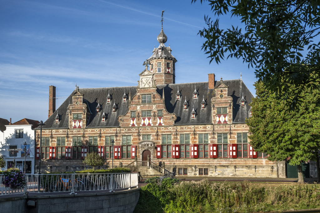 Historisch pand in Middelburg met classicistische architectuur en rode en witte geveldecoraties, gelegen aan de rivier, onder een blauwe hemel.