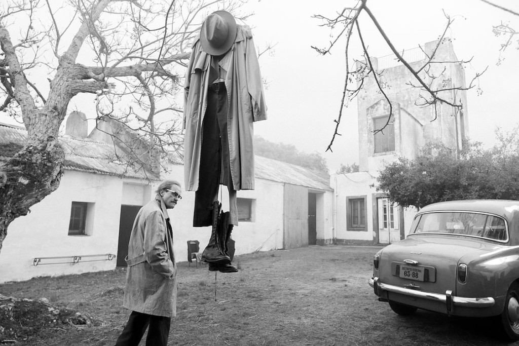 Droogstaand en surrealistisch beeld met man die naar ingedroogde jas en hoed op kledinghanger onder een boom kijkt, naast vintage auto, straat en kerktoren in de achtergrond, zwart-wit.