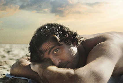 Een jonge man ligt op het strand, ontspannen en kijkend naar de camera, met een luchtige, zomerse sfeer die uitnodigt tot film en entertainment bij Cinema Middelburg.