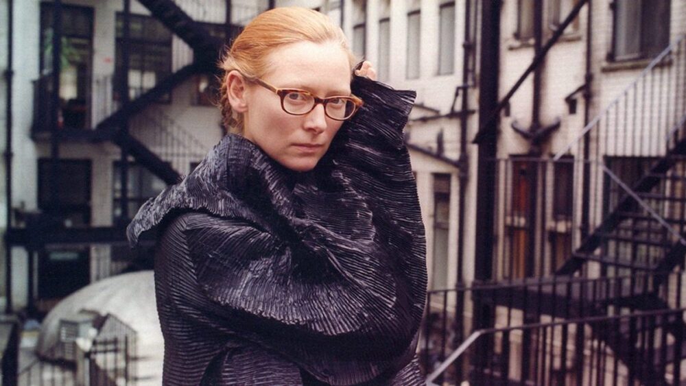 Zwarte vrouw met glazen in een stadstuin, modevolle jas, urban achtergrond, straatfoto, actueel, stijlvol, straatmode, vrouwelijke fashion, stedelijk leven, casual, trendy.