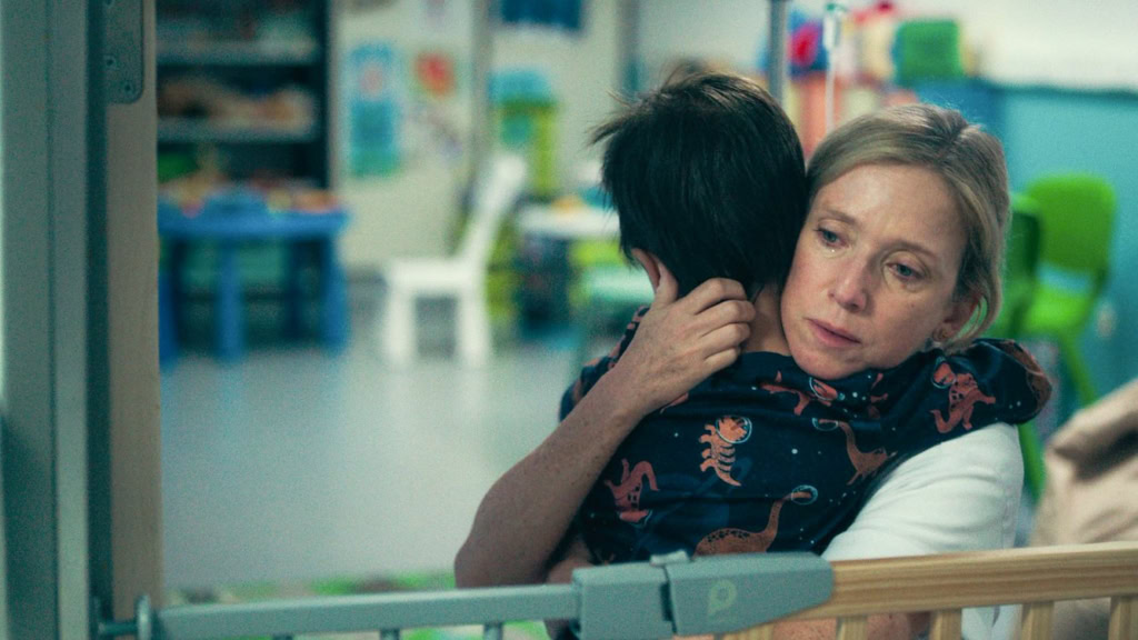 Een vrouw omarmt een kind in een verzorgingshuis, met een zachte uitdrukking van verdriet en troost. De setting is kinderopvang of medische omgeving, met kleurrijke achtergrondelementen en een intiem moment van verbondenheid.