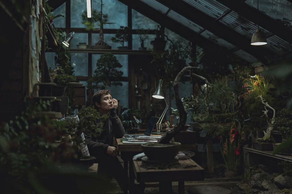 Kunstzinnige foto van een vrouw die in een botanische kas zit, omringd door planten en bomen, met een laptop en boeken, onder sfeervol licht, passend bij de film 'No Other Choice' en de sfeer van Cine.