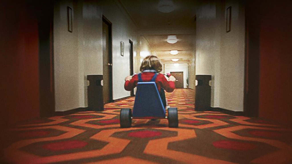 Kleine jongen op loopauto rijdt door een hotelgang met een kleurrijk tapijt, onder de titel "The Shining (45th Anniversary)".