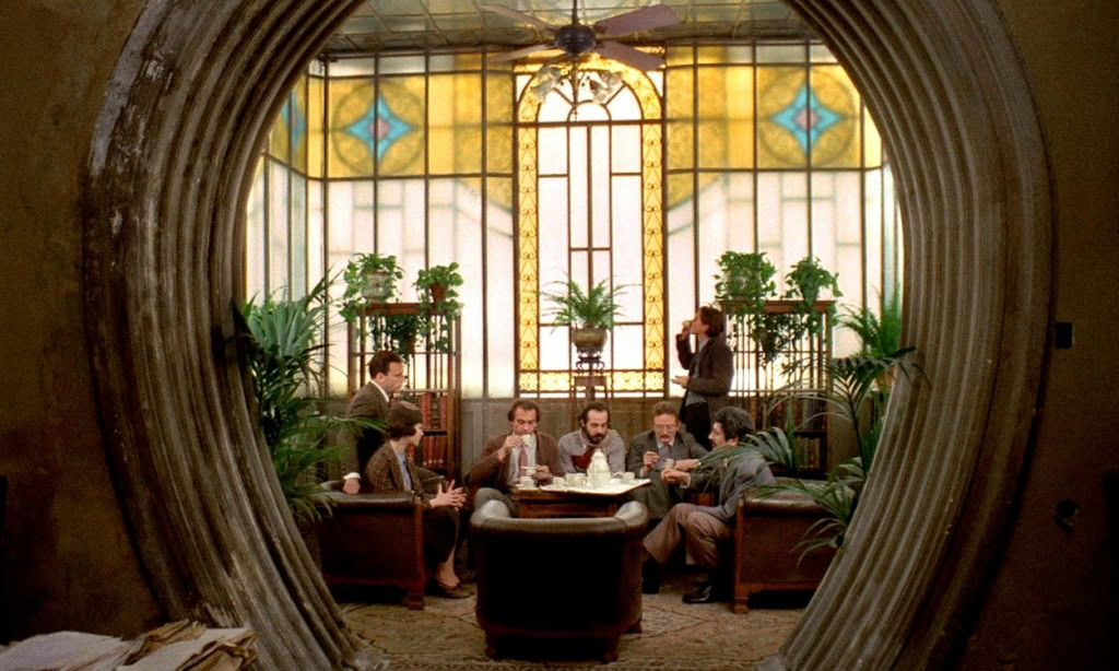 Gezicht op een elegante, historische woonkamer met grote stained glass ramen, waarin een groep mannen in formele kleding samenkomt, met een filmthema dat past bij de restauratie van Il Conformista.
