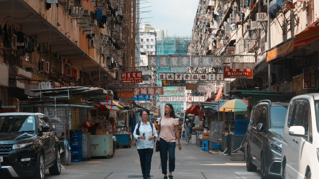 Gezicht op een drukke straat in Hong Kong met winkels en marktkramen, onder een overdekte markt met Chinese tekens en reclameborden, met twee vrouwen die wandelen en genieten van de omgeving.