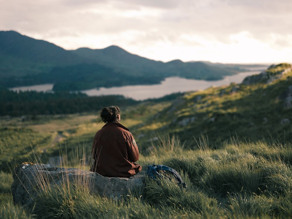 Een persoon zit op een rots in een uitgestrekt landschap met groene heuvels, bergen en een meer op de achtergrond, genietend van de rust en de natuurlijke omgeving.
