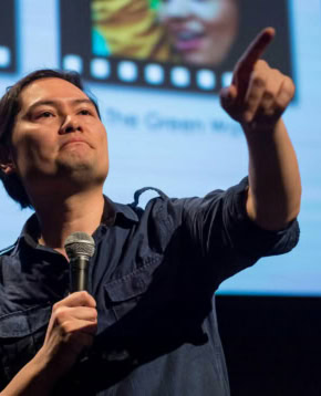 Een spreker tijdens een filmcollege presentatie in Cinema Middelburg, met een microfoon in de hand en een scherm met filmbeelden op de achtergrond.