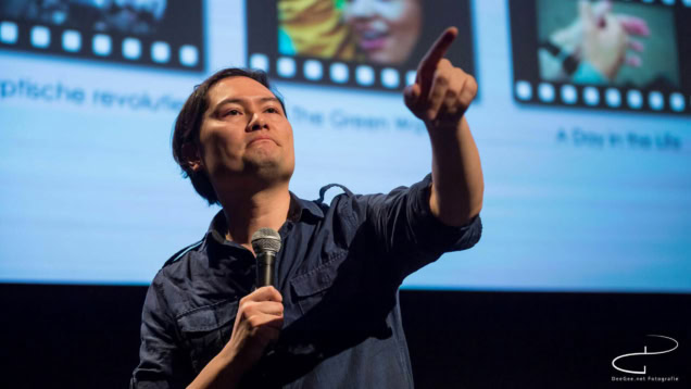 Een spreker tijdens een filmcollege presentatie in Cinema Middelburg, met een microfoon in de hand en een scherm met filmbeelden op de achtergrond.