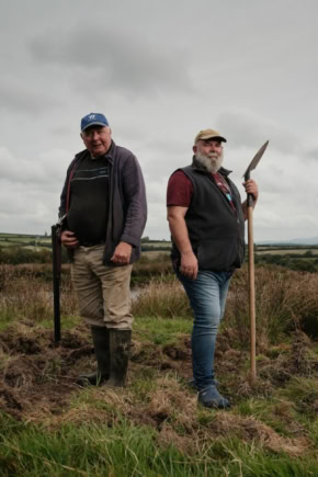 Twee mannen gekleed als filmhelden staan in een open veld met een landschap op de achtergrond, symboliserend de film "Movies That Matter On Tour: Derek vs Derek".