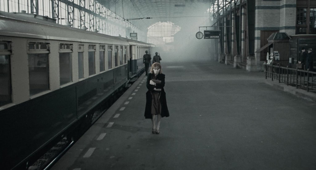 Een vrouw staat alleen op een verlaten station, omringd door oude treinen en industriële gebouwen, met een mysterieuze sfeer die past bij de restauratie van de film 'Het meisje met het rode haar'.