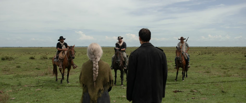 Een scène uit de film 'Horizonte' met vier cowboys op paarden en twee toeschouwers in een uitgestrekt grasland onder een bewolkte hemel, geschikt voor filmvertoningen en culturele evenementen.