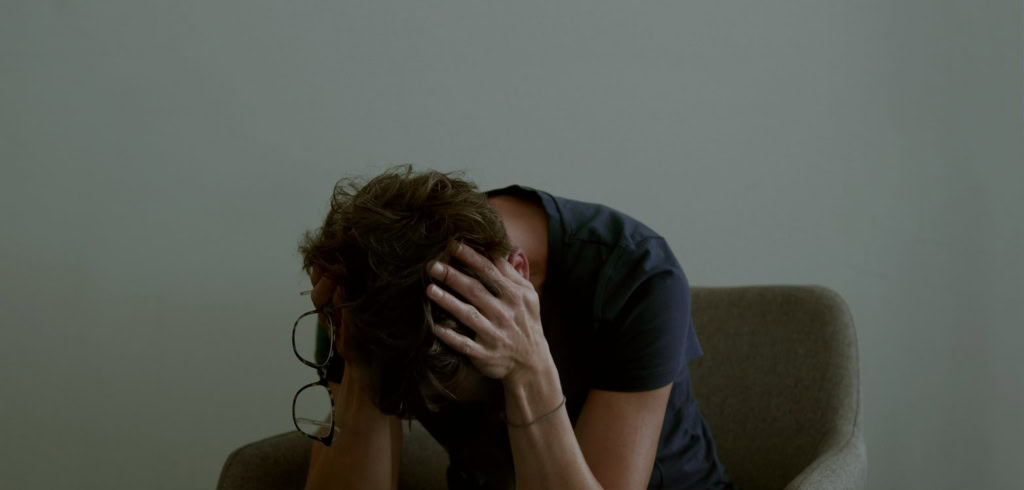 Een persoon met kort haar zit op een bank, met hoofd in handen, uitdrukking van verdriet of frustratie, symboliseert emotionele strijd en persoonlijke reflectie, passend bij de titel "Mijn woord tegen.