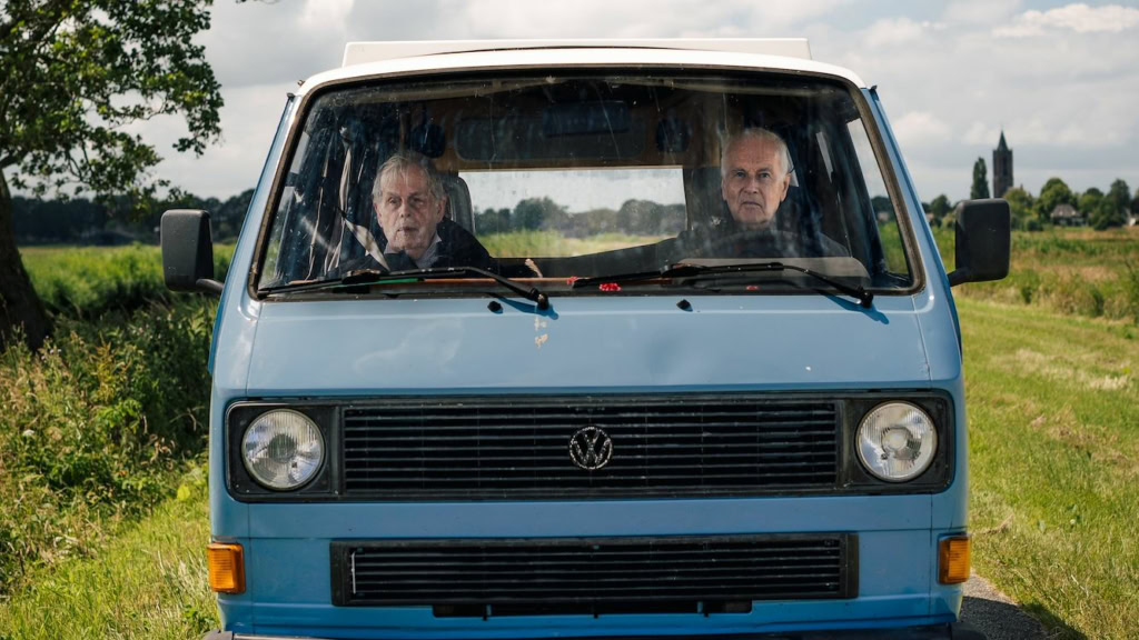 Twee mannen zitten in een blauwe bus, omringd door groene velden en een heldere hemel, met een landelijke achtergrond. Perfect voor liefhebbers van Nederlandse films en filmtheaterervaringen bij Cinem.