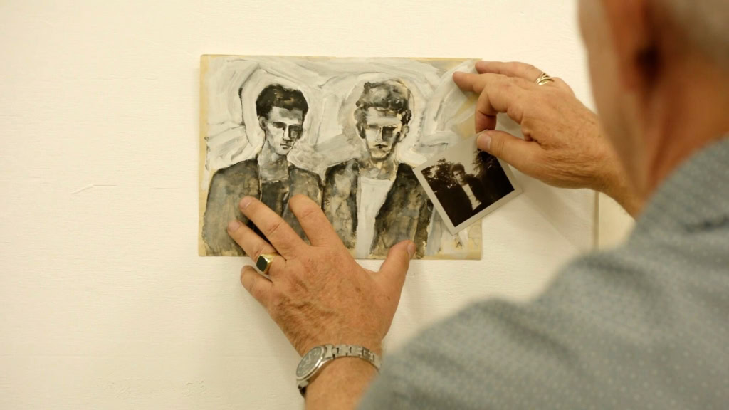 Een persoon die een zwart-wit schilderij vasthoudt van twee mannen, met een foto in de hand, in een kunstgalerie.