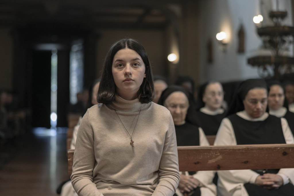 Jong meisje zit stil in de kerk tijdens de voorstelling 'Los Domingos', omringd door nonnen, met een serene en contemplatieve sfeer, perfect voor een film over religieuze of culturele thema's.