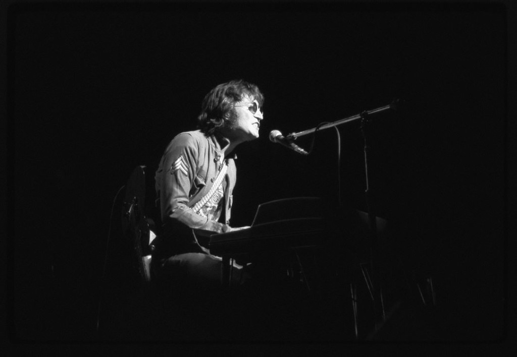 Zwarte en witte foto van John Lennon tijdens een live optreden in New York City, zittend achter een piano en zingend in een donkere setting.