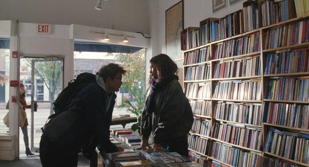 Twee mensen praten in een boekwinkel met veel boeken op de achtergrond, voor een film getiteld "De Drama" bij Cinema Middelburg.