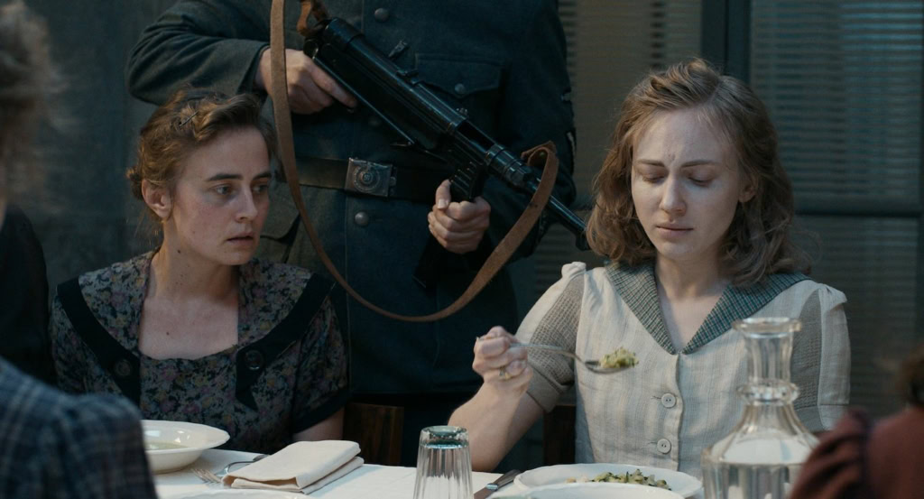 De afbeelding toont een scène uit de film "De Tasters" met twee vrouwen die aan een diner deelnemen, terwijl een gewapende man op de achtergrond staat.