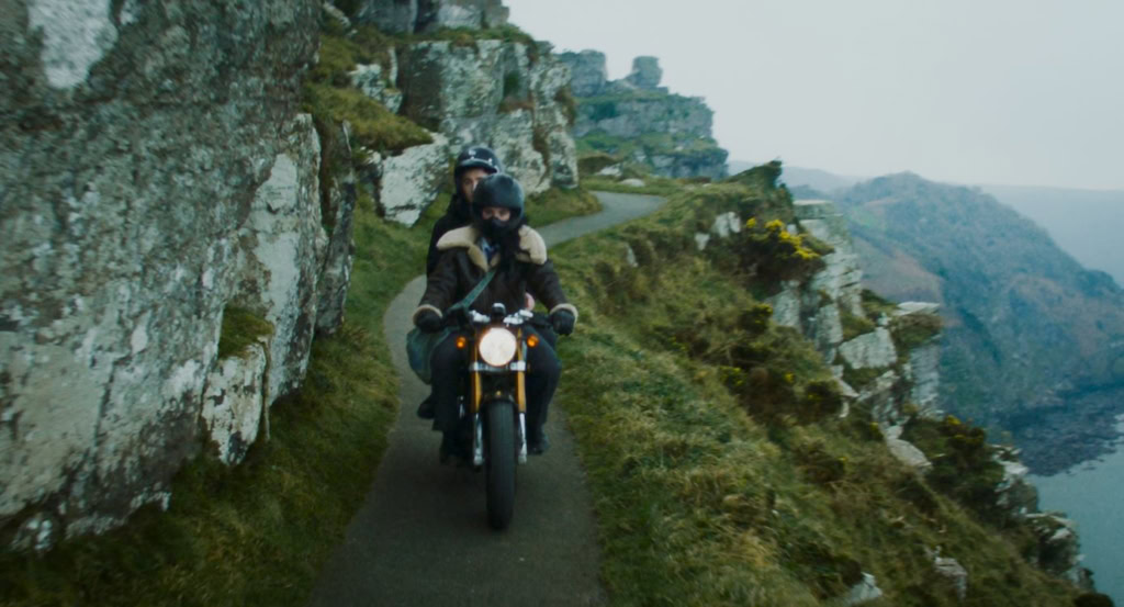 Twee motorrijders rijden langs een smal pad langs de kliffen met uitzicht op de zee, omgeven door groene heuvels en rotsformaties, onder een bewolkte hemel.