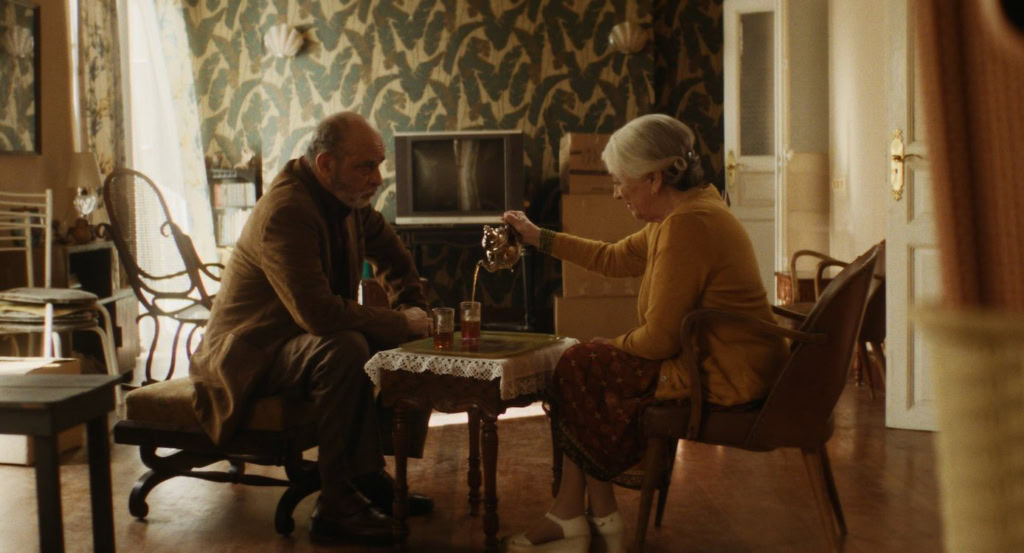 Gezellige filmavond met twee oudere mensen die samen in een woonkamer zitten, genieten van een drankje en een film kijken in een knusse, warme sfeer.