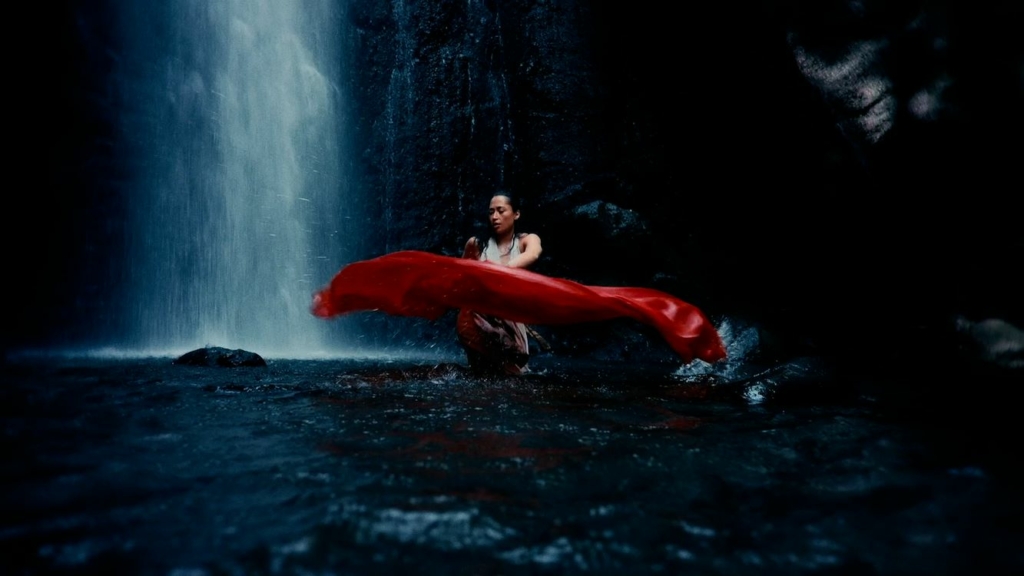 Een vrouw staat in een donkere, natuurlijke waterval met een rode doek, omgeven door rotsen en stromend water, perfect voor een beeld dat kracht en rust uitstraalt.