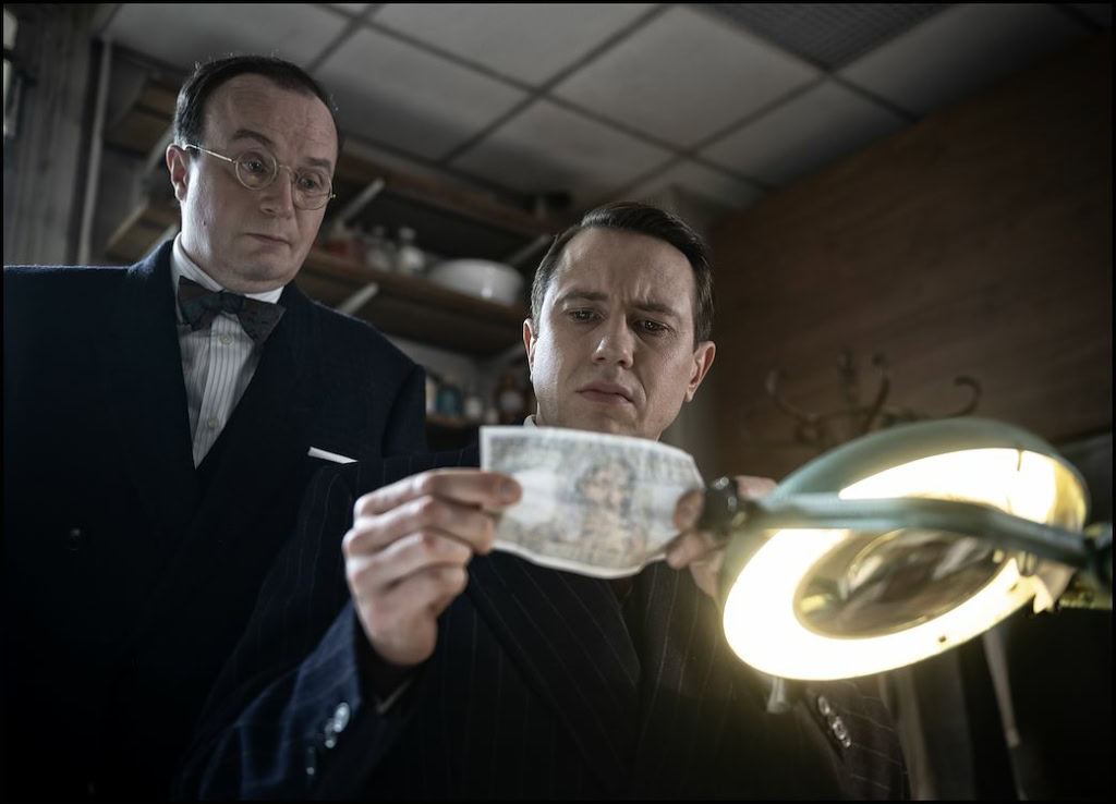 Een scène uit een film met twee mannen in formele kleding, één die een stuk papier of een document onderzoekt onder een lamp, in een filmstudio of kantooromgeving, geschikt voor een bioscoopwebsite.