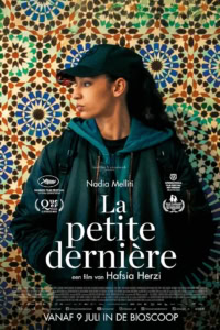 Afbeelding van een jonge vrouw in een zwart jasje en blauwe hoodie voor een kleurrijk mozaïekmuur, promotieposter van de film La Petite Dernière.