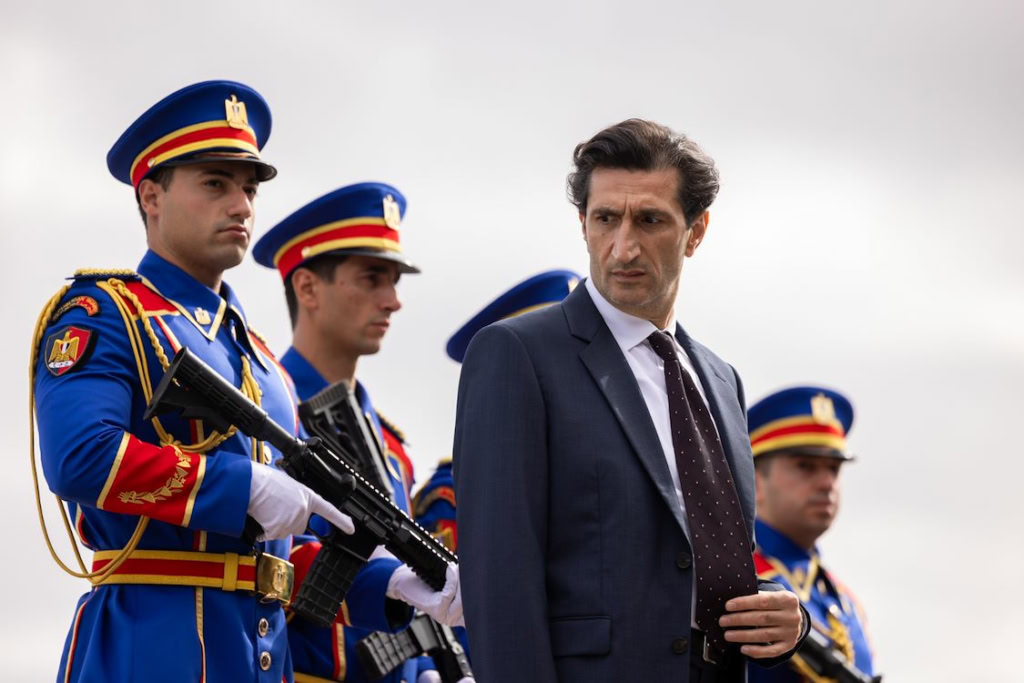 Mannelijke officier in pak met vier soldaten in blauwe uniformen en helmen, militaire parade of ceremoniële gebeurtenis, dramatische sfeer, buitenlocatie onder bewolkte hemel.