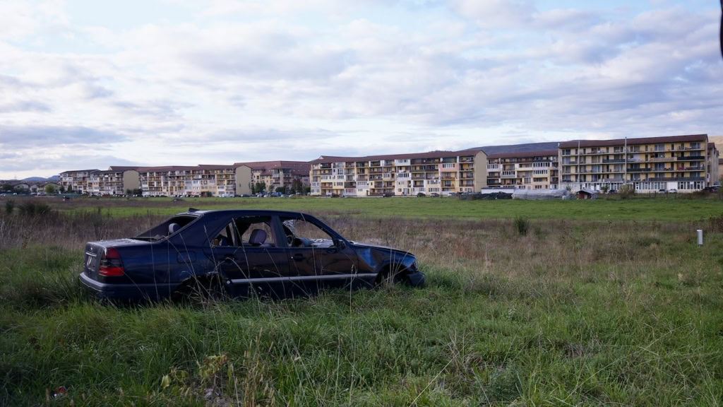 Verlaten zwarte auto op een grasveld met moderne appartementen op de achtergrond, onder een bewolkte hemel, nabij Cinema Middelburg.