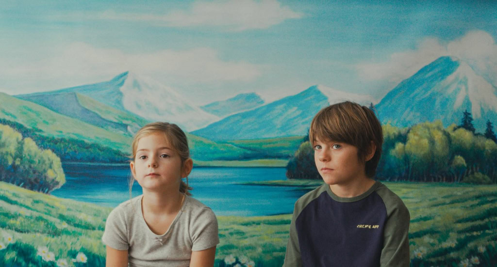 Twee kinderen zitten voor een grote muur met een schilderij van bergen, een meer en groene heuvels, in een filmzaal of theater.