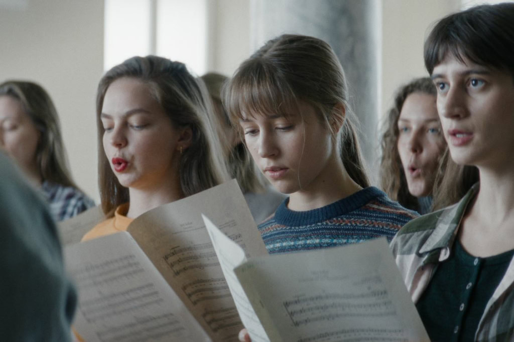 Groep jonge vrouwen die samen zingen en een liedje oefenen in een filmtheater, met focus op zang en samenwerking.