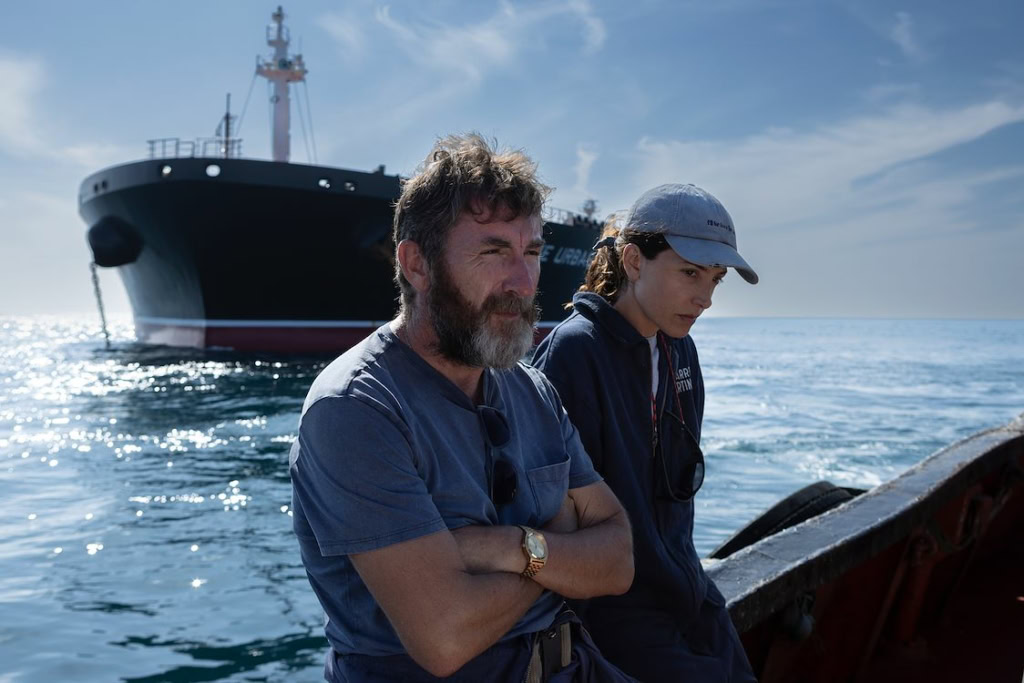 Twee mensen op een boot met een groot schip op de achtergrond, zee en blauwe lucht, filmische sfeer.
