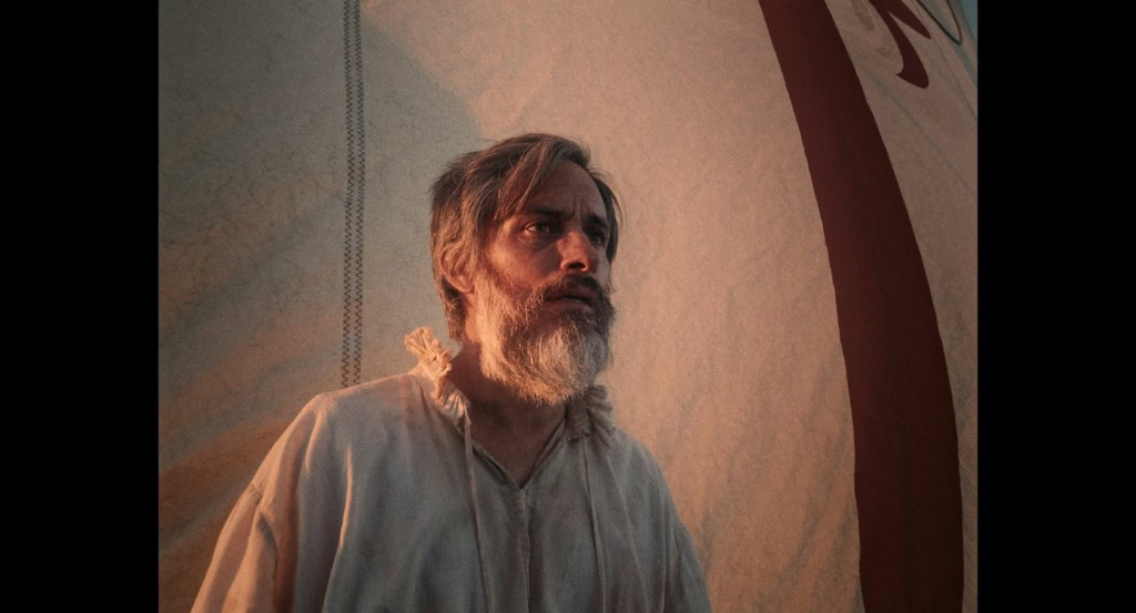 Man met lang haar en baard in een tent, filmset of decor, met een dramatische sfeer, voor een film of theaterproductie.