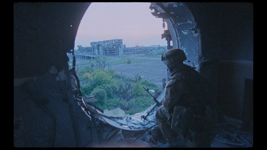 Soldaat kijkt uit het raam van een vliegtuig met uitzicht op een verstedelijkt landschap en groene gebieden, binnen een militaire setting.