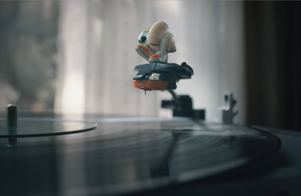 Close-up van een vinylplaat met een speelgoedfiguur van Marcel de Schelp die op een platenspeler draait, in een sfeervolle kamer met natuurlijk licht.