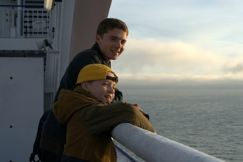 Twee jonge mannen genieten van het uitzicht vanaf het dek van een schip, met de zee en een ondergaande zon op de achtergrond, tijdens een filmvertoning.