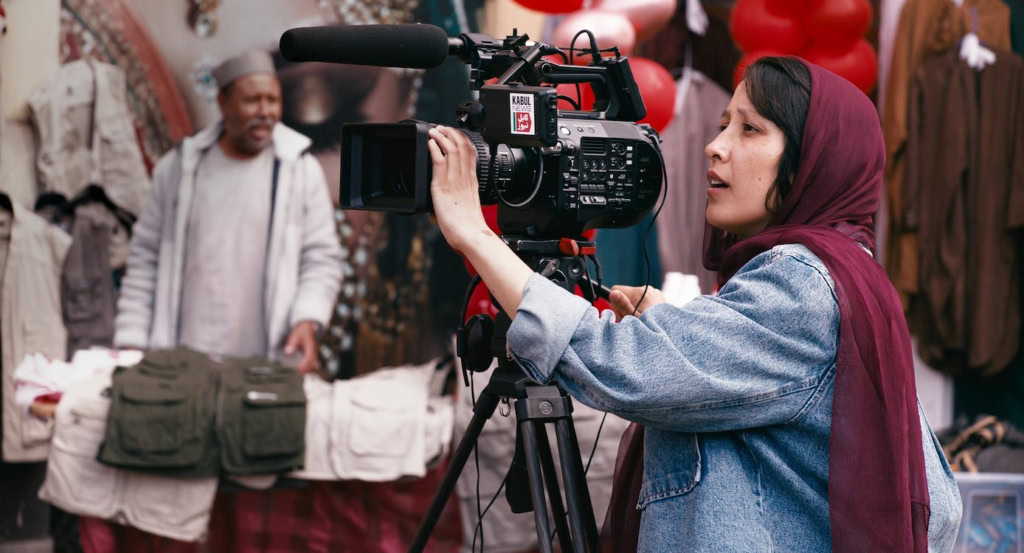 Vrouw met hoofddoek filmt met professionele camera op markt in Middelburg, kleurrijke achtergrond met mensen en kledingstalletjes.