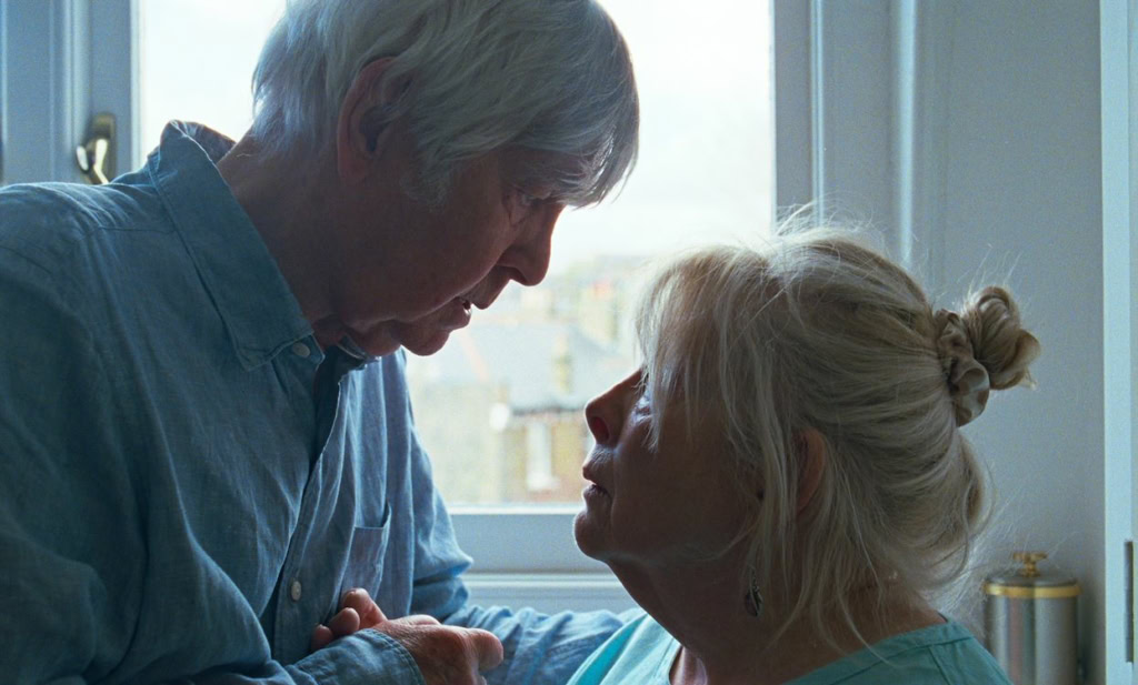 Een oudere man en een oudere vrouw kijken intens naar elkaar in een huiselijke setting, met een raam op de achtergrond dat natuurlijk licht binnenlaat.