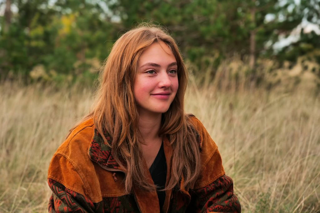 Gezicht van een jonge vrouw met lang, lichtbruin haar, zittend in een veld met gras en bomen op de achtergrond, genietend van de natuur.