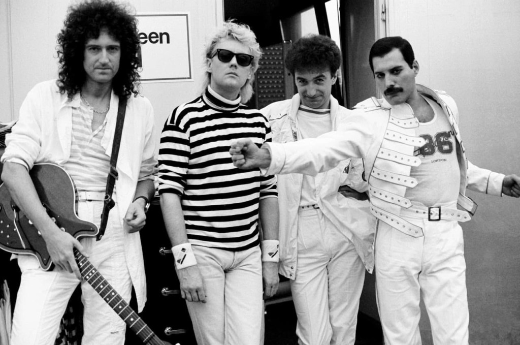 De band Queen Budapest poseert in zwart-wit foto, met een gitarist, zanger en andere bandleden in opvallende outfits, klaar voor een optreden.