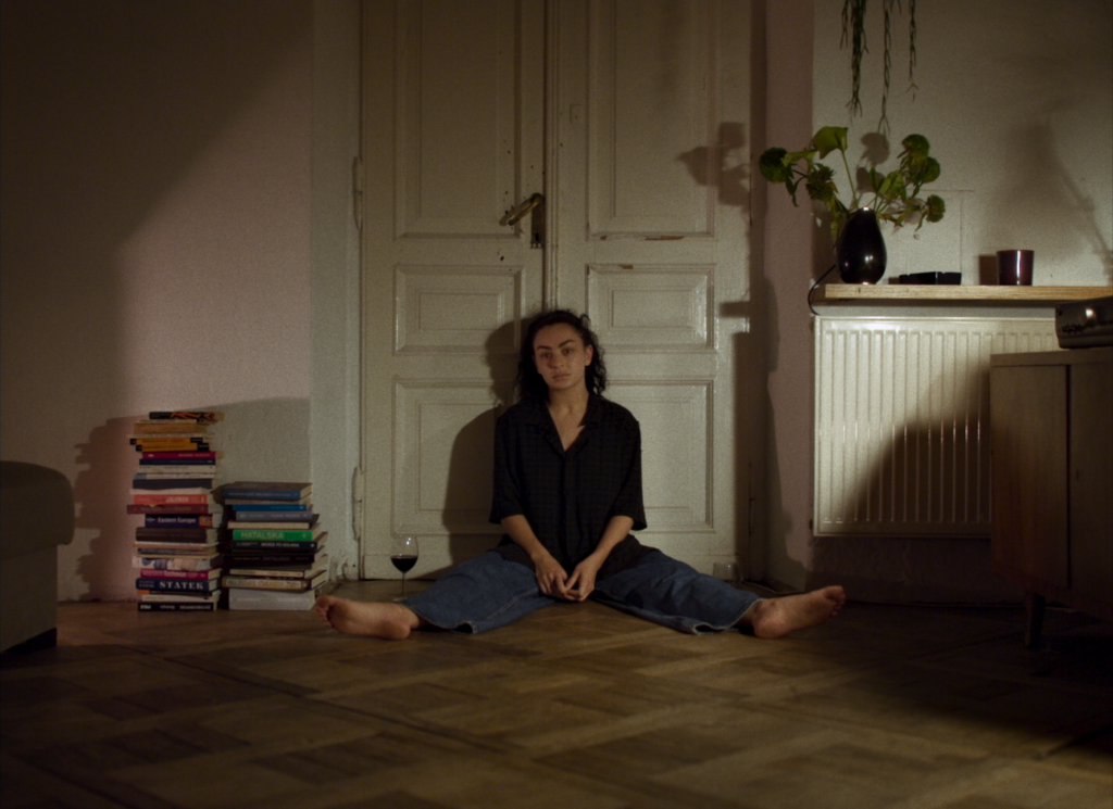 Persoon zit op de vloer in een kamer met boeken, planten en een radiator, met een rustige en introspectieve sfeer.
