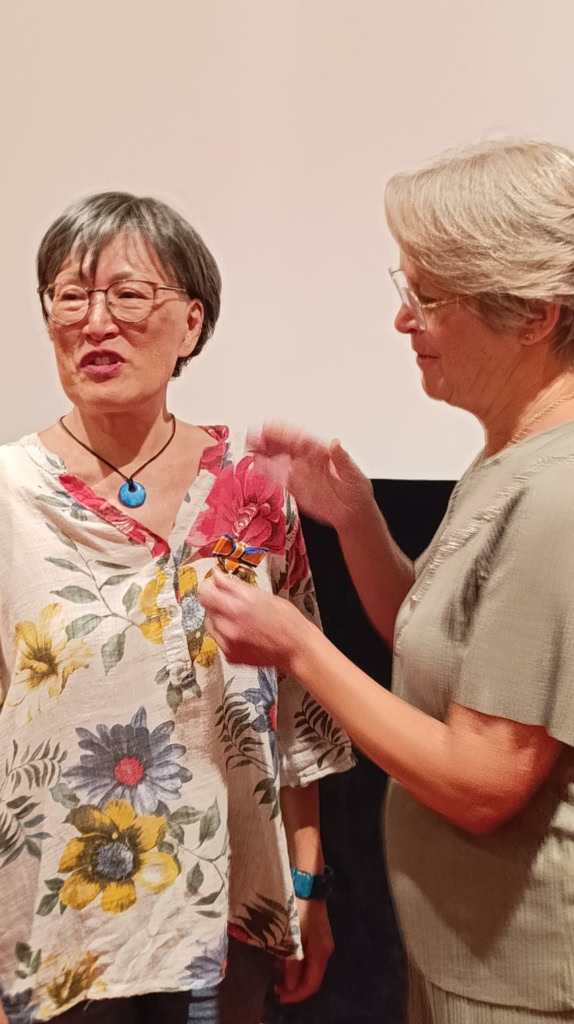 Hedy Tjoeng krijgt een Koninklijke onderscheiding tijdens een ceremonie, omringd door andere genodigden, in een formele setting.