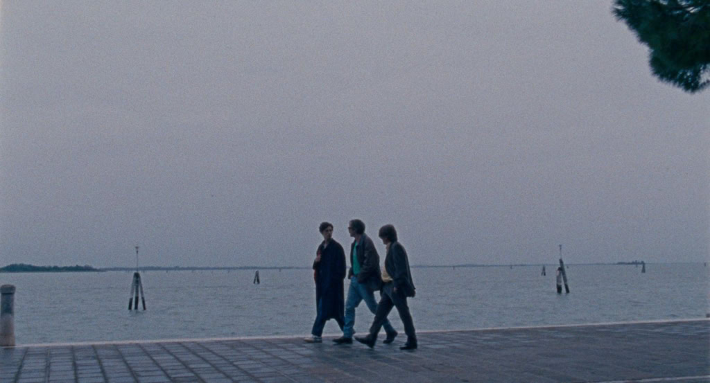Drie mensen wandelen langs de waterkant bij zonsondergang, met een rustige zee en een heldere hemel op de achtergrond.