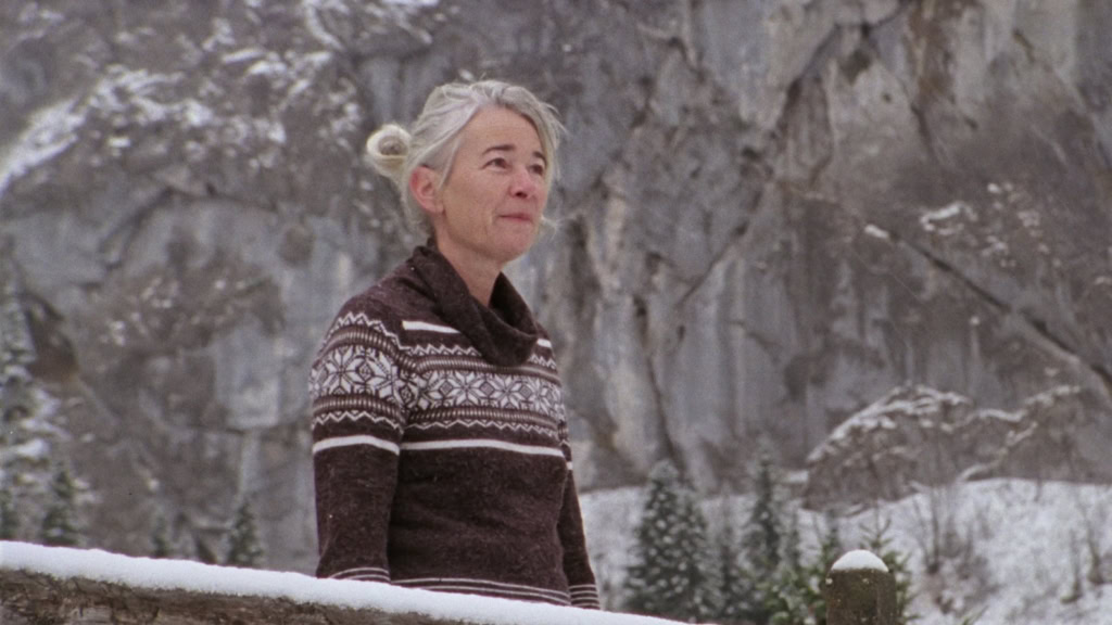 Oudere vrouw met grijs haar en een gebreide trui kijkt uit over een besneeuwd berglandschap, met een serene uitdrukking.