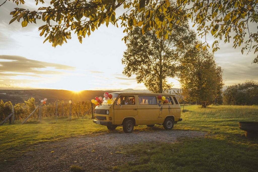 Een gele bus met ballonnen geparkeerd in een groene veld bij zonsondergang, omgeven door bomen en een rustige landelijke omgeving.